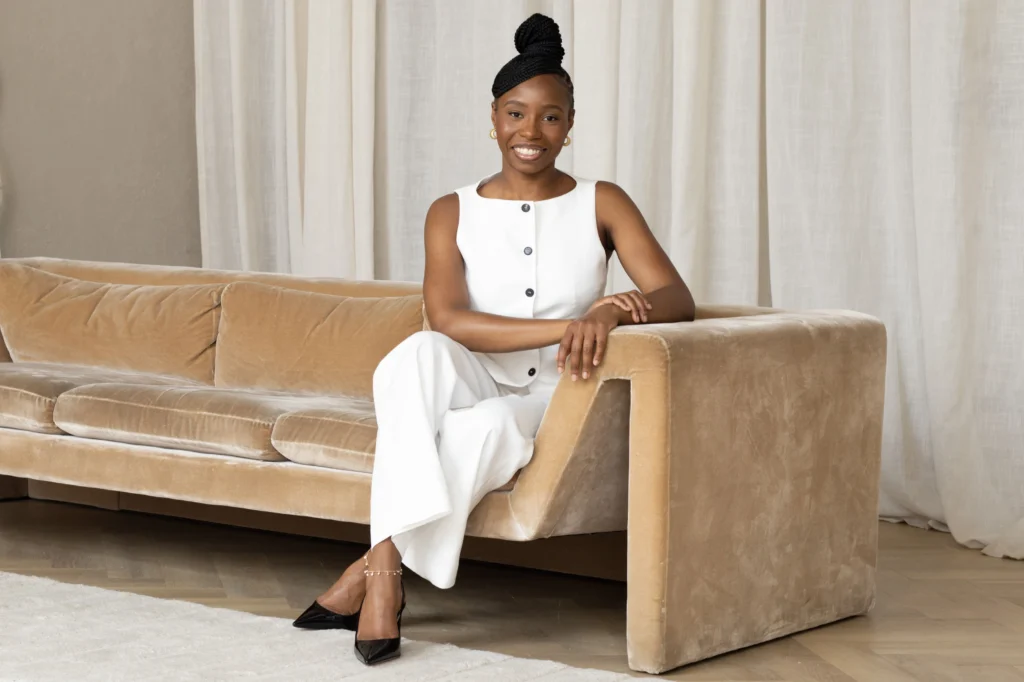 Smiling Cashala Eugene in white outfit seated on beige velvet sofa